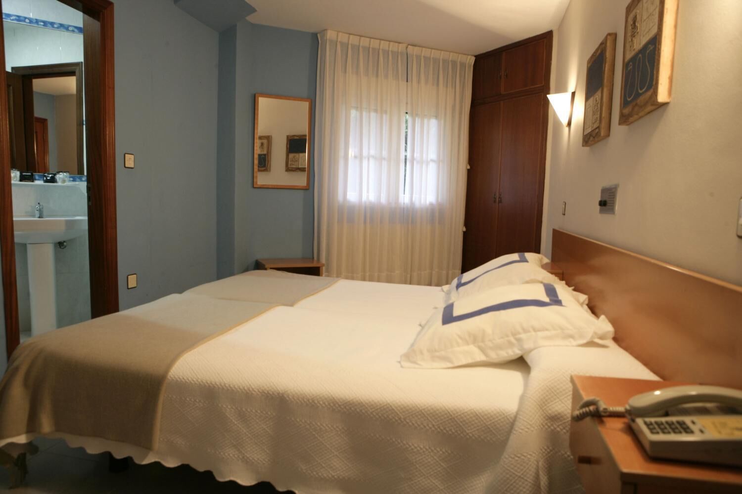 A hotel room with a bed, a bedside telephone, a mirror, a window with curtains, and a wooden wardrobe.