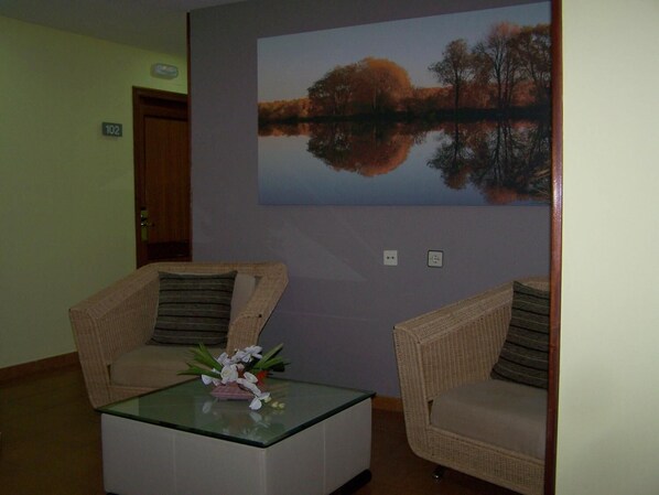 Una habitación con dos sillones de mimbre, una mesita de centro de vidrio y un arreglo floral, además de un cuadro grande que representa un lago.