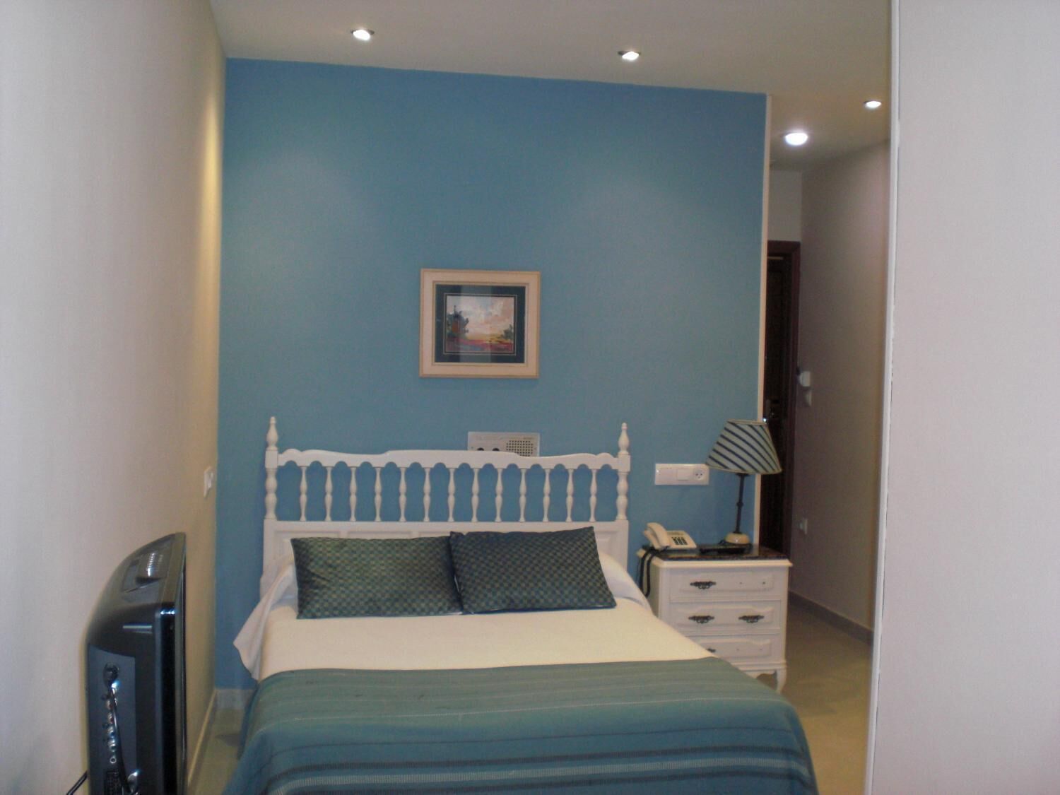 Un dormitorio con cama, mesita de noche con lámpara, teléfono y televisor.