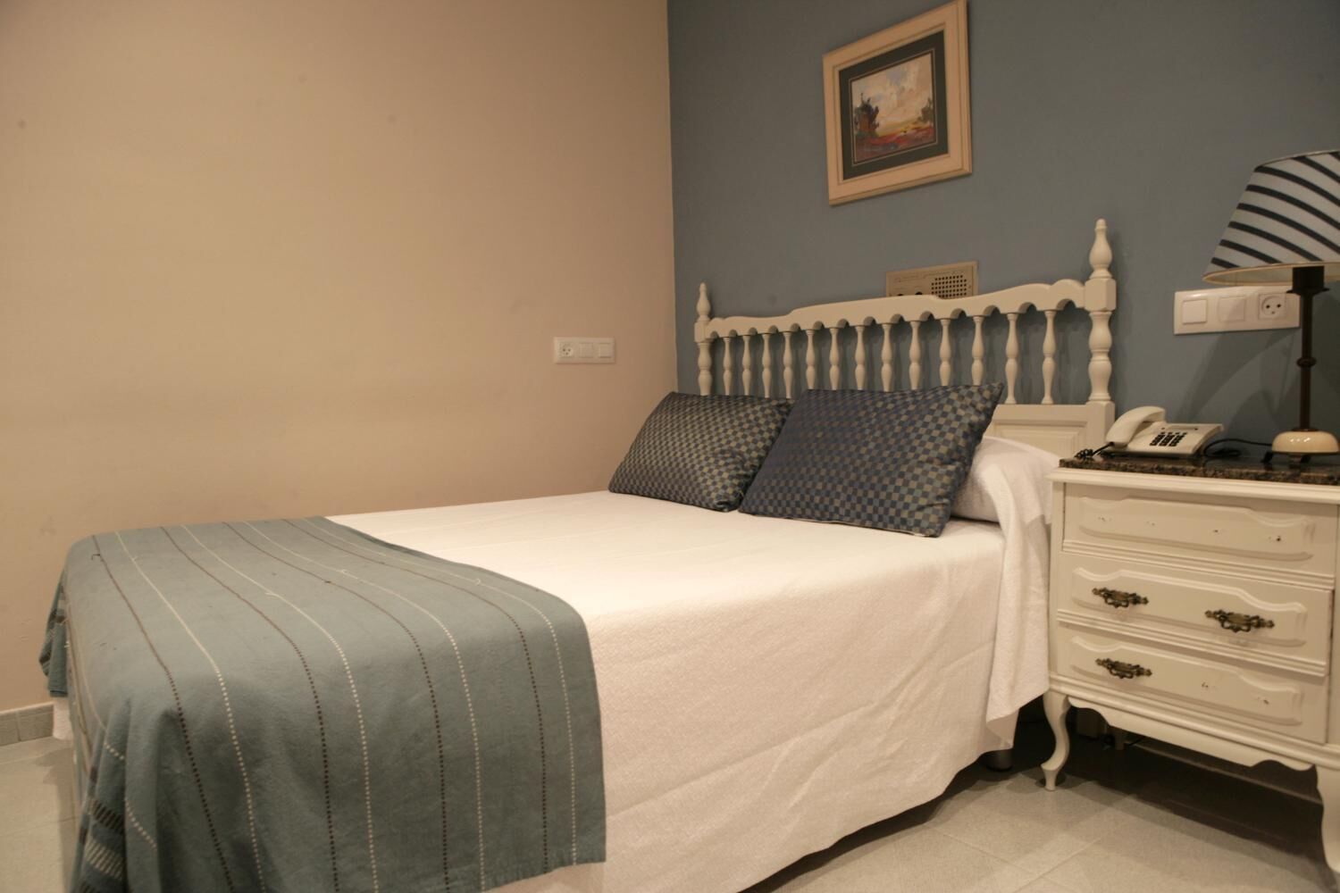 A neatly made bed with a striped blanket and two pillows, a bedside table with a lamp and a phone, and a painting on the wall.