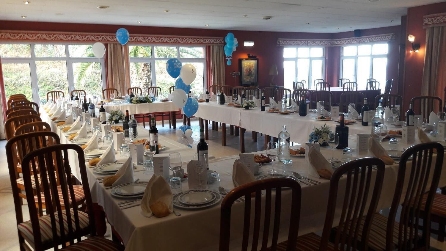 Un salón de banquetes con largas mesas dispuestas para un evento formal, con manteles blancos, sillas ordenadas y un gran ventanal que proporciona luz natural.
