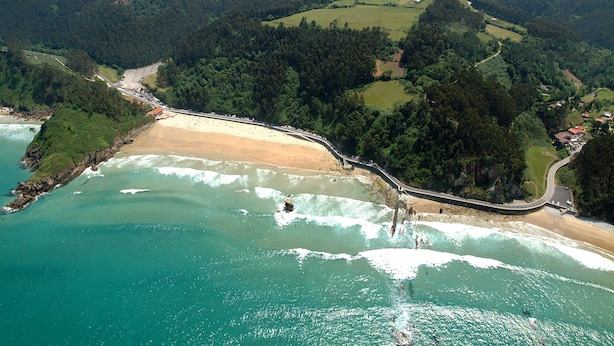 Vista aérea de una zona costera con playa, carretera y colinas boscosas circundantes.