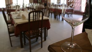 Un comedor con mesas dispuestas para comidas, una copa de vino sobre una mesa, y vista del cuarto con ventanas y un cuadro en la pared.
