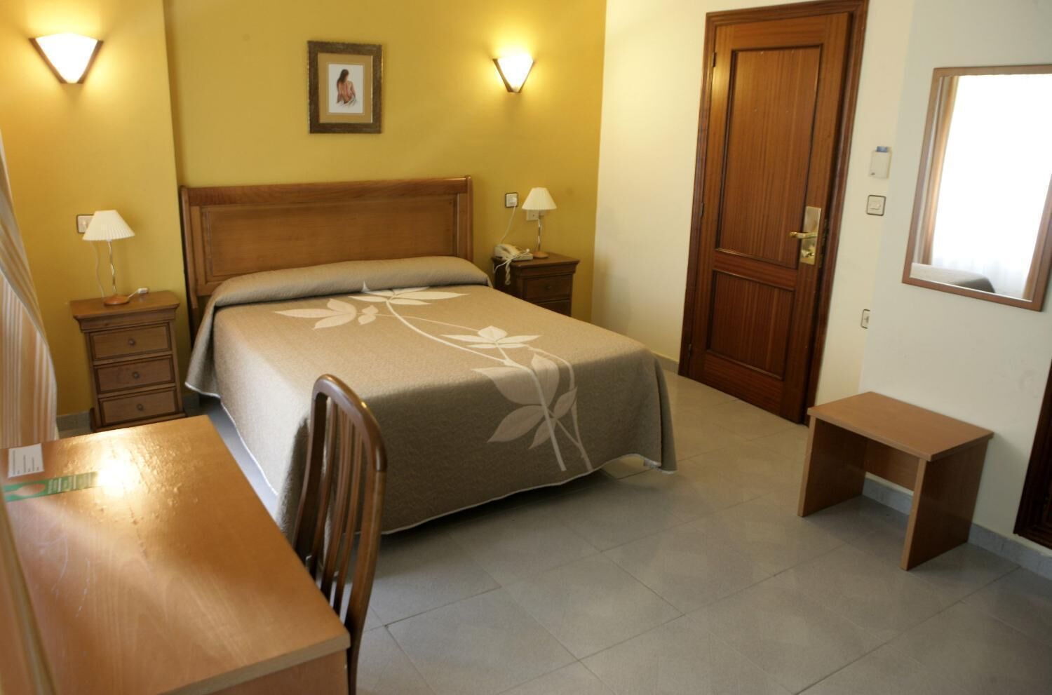 Habitación de hotel con cama, escritorio, silla, dos mesitas de noche con lámparas, puerta de madera y un cuadro en la pared.