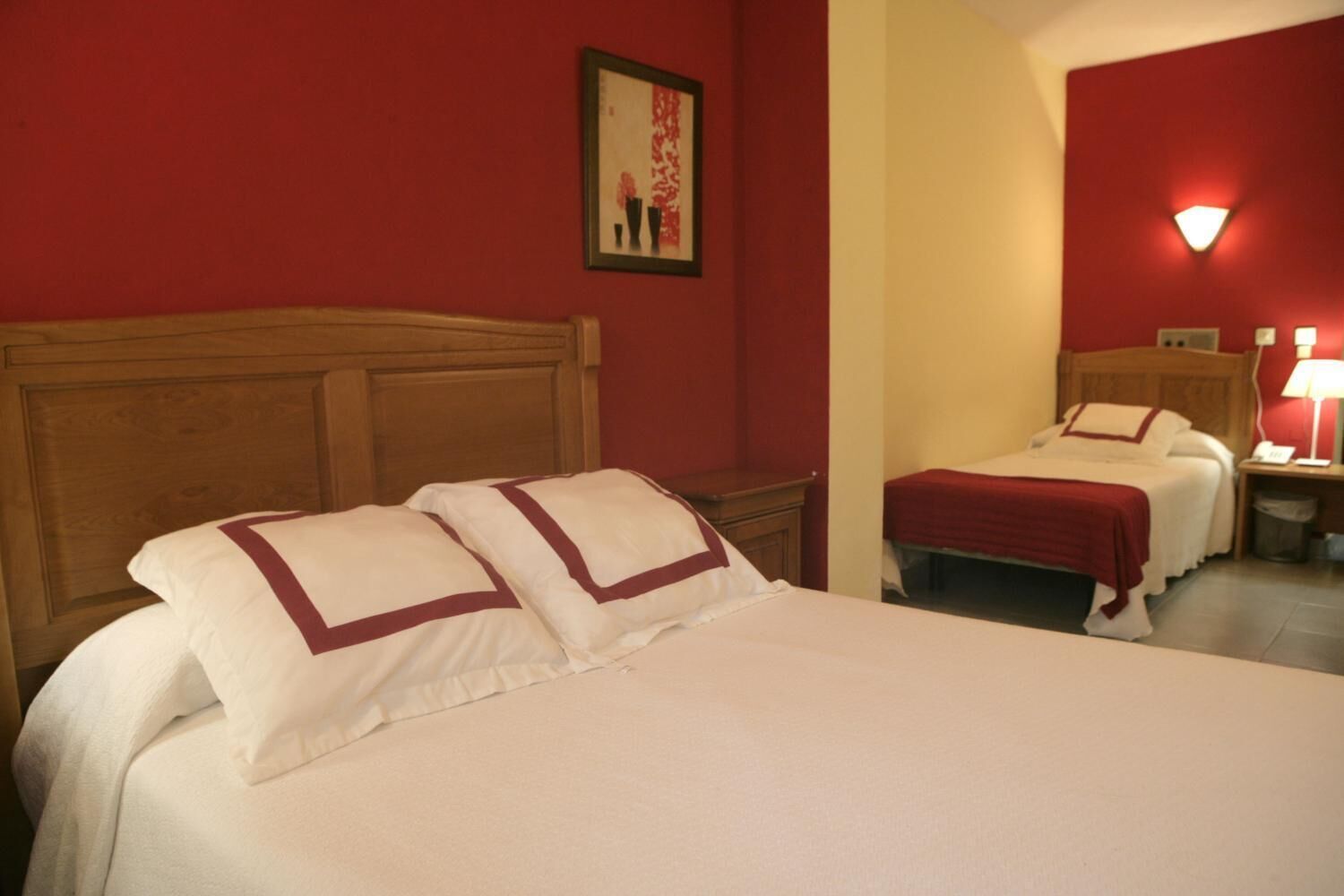 Habitació d'hotel amb dos llits, un capçal de fusta, una paret vermella i un quadre amb marc.