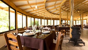 A restaurant with wooden beams, large windows, and a central wood stove. Tables are set with plates, glasses, and cutlery. - Toucanet Lodge (Copey)
