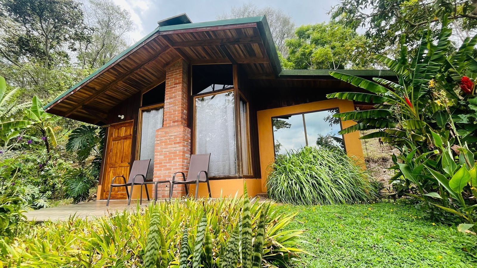 Une maison moderne avec une cheminée en briques, de grandes fenêtres et une véranda en bois avec des chaises.