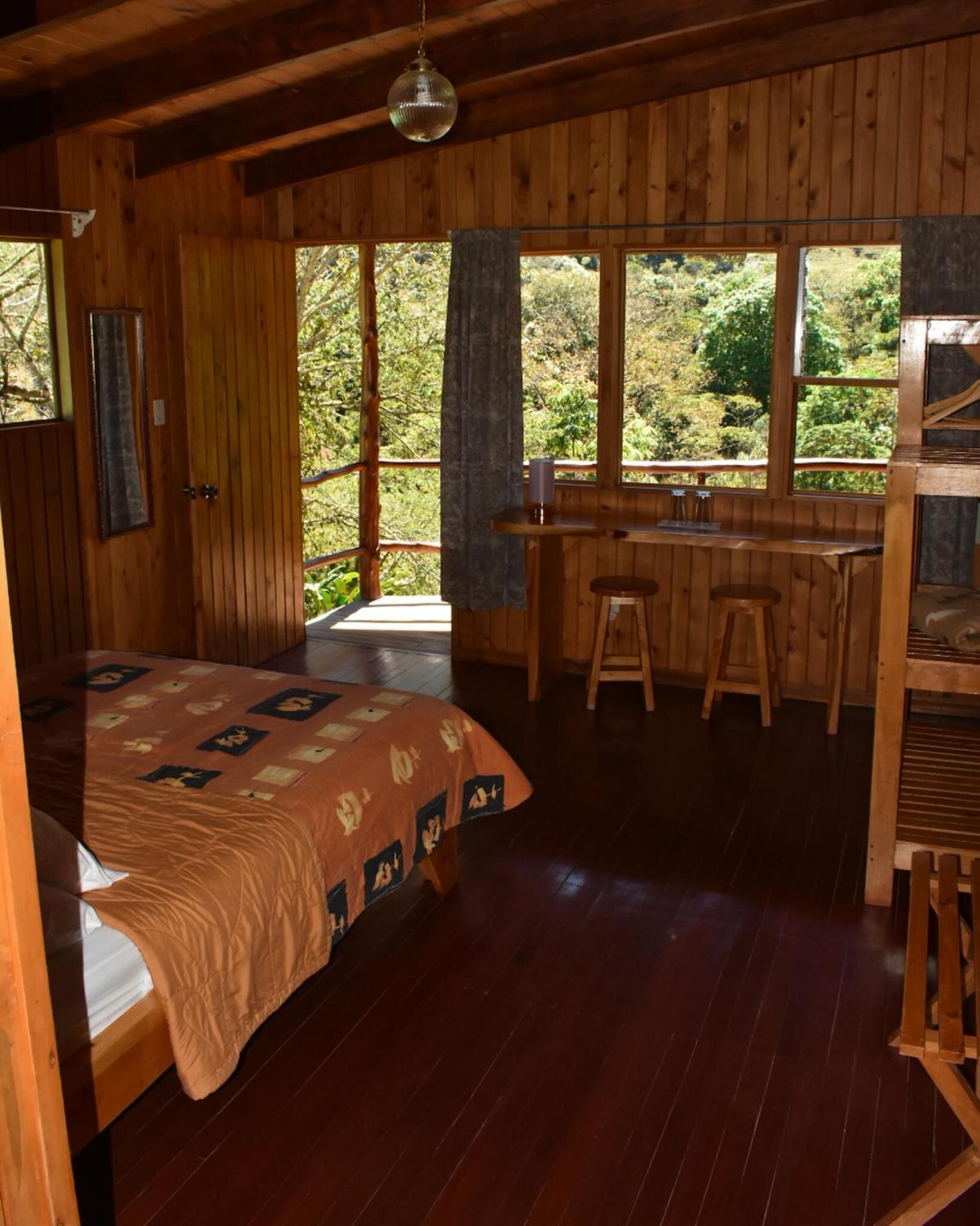 Una cabaña de madera con una cama, una zona de comedor con taburetes y grandes ventanales que dan al bosque.