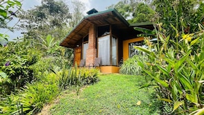 A small, modern house with a brick exterior and large windows, surrounded by lush greenery and a well-maintained garden. - Toucanet Lodge (Copey)