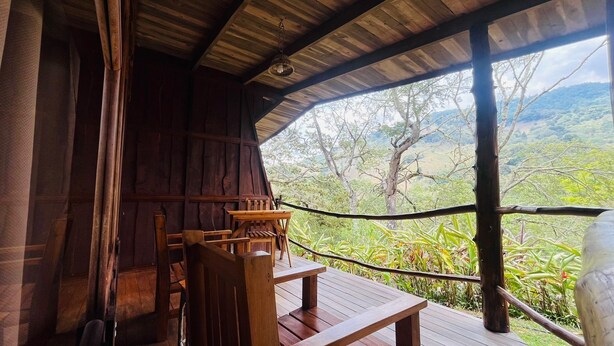 A wooden cabin with a table and chairs, a hanging light fixture, and a view of a lush landscape. - Toucanet Lodge (Copey)