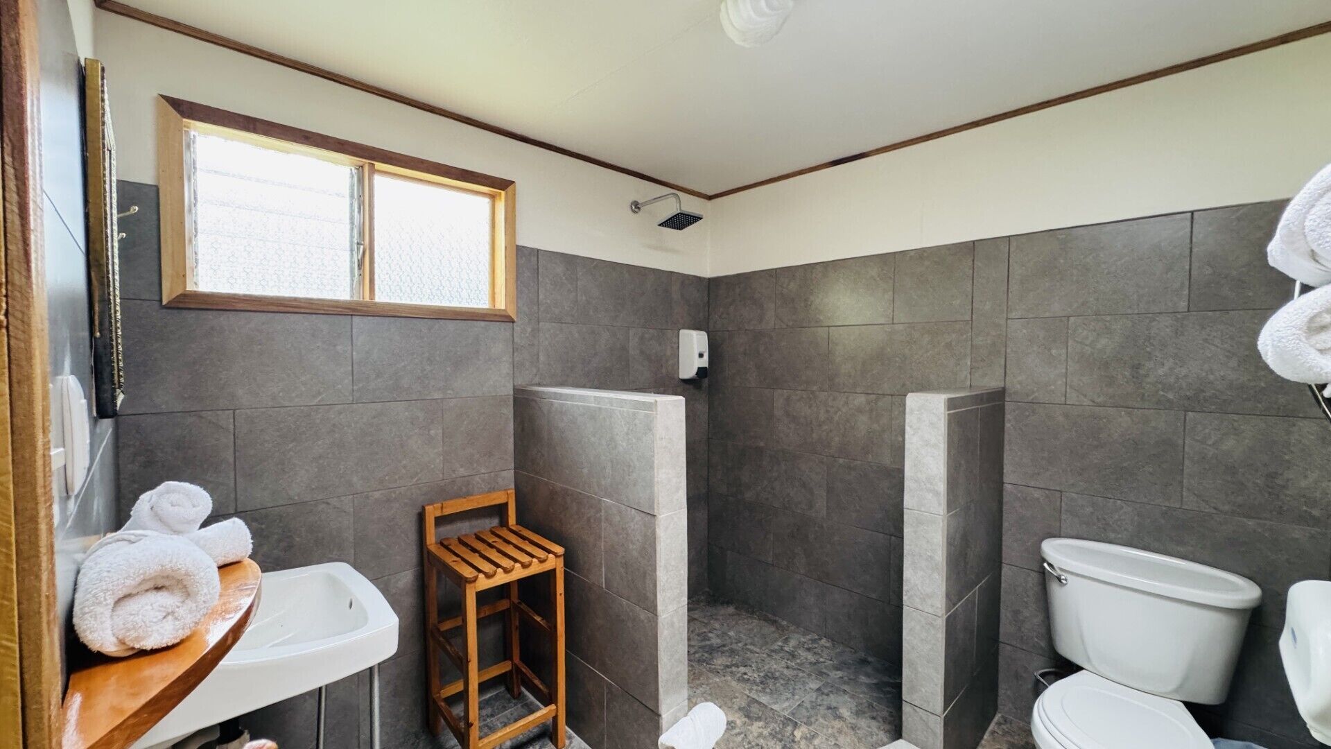 Baño con ducha, inodoro, lavabo y un pequeño taburete de madera.