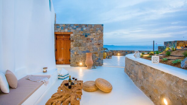 Un espace extérieur ensoleillé, aménagé avec des sièges, un mur en pierre, une porte en bois et une vue sur la mer.