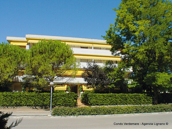 A multi-story building with a yellow facade, surrounded by greenery and a clear blue sky.
