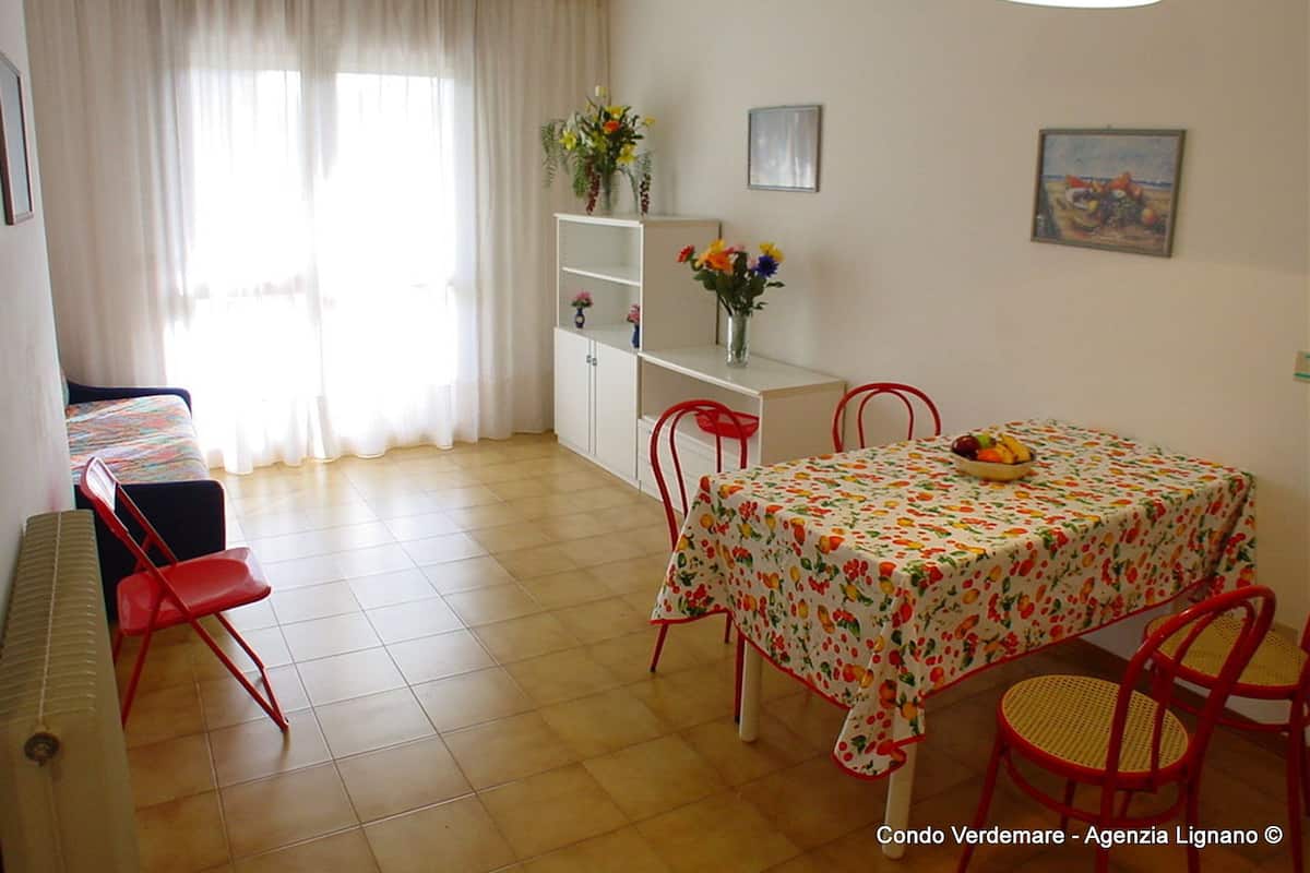 A dining area with a table covered in a fruit-patterned tablecloth, surrounded by red chairs. There is a white cabinet with shelves, a vase with flowers, and a painting on the wall.