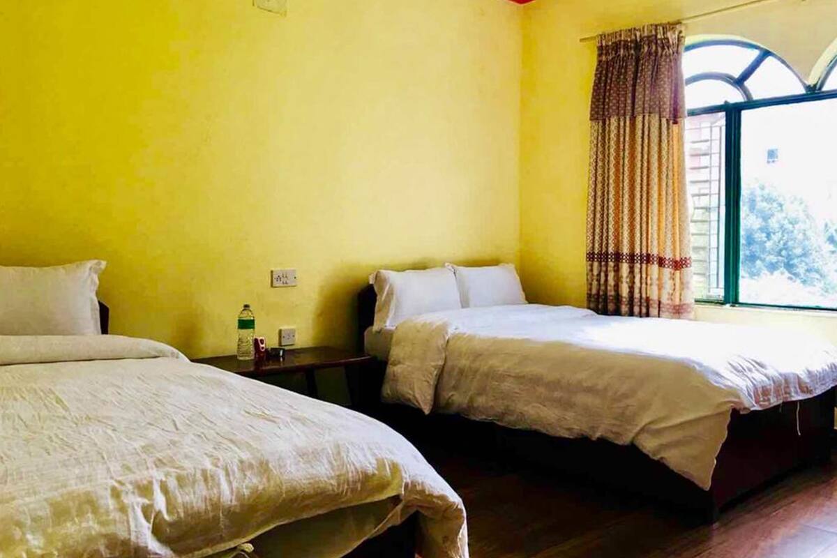 A room with two beds, a window with curtains, and a small table with a bottle and a cup.