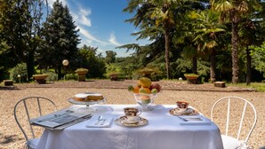 Una mesa al aire libre con mantel blanco, rodeada de sillas, un frutero y platos de comida.