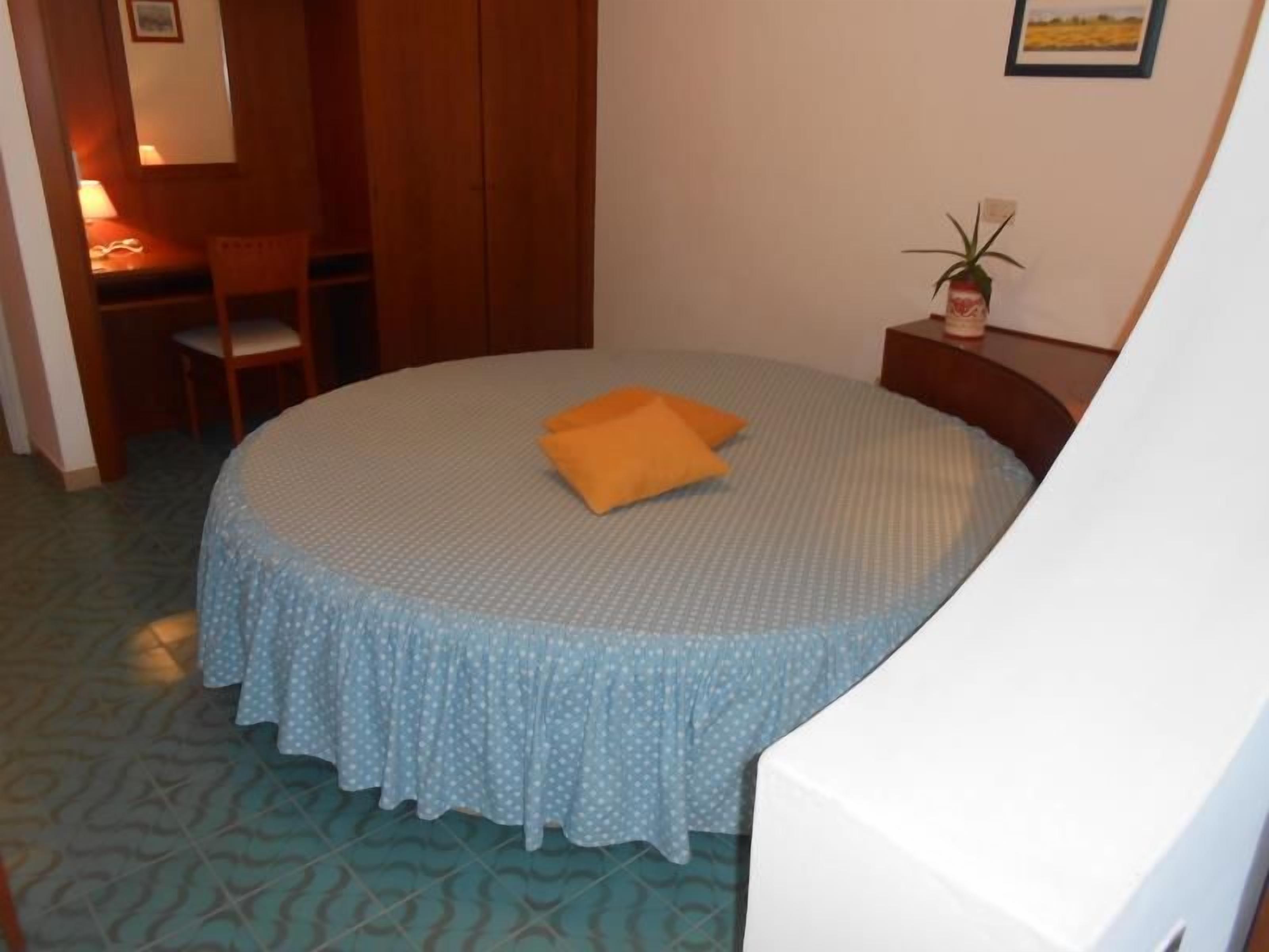 Una cama redonda con colcha celeste con lunares y almohada naranja, una mesita de noche de madera con una planta, y una silla en una habitación con alfombra estampada y paredes de madera.