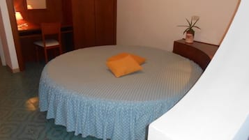 Una cama redonda con colcha celeste con lunares y almohada naranja, una mesita de noche de madera con una planta, y una silla en una habitación con alfombra estampada y paredes de madera.