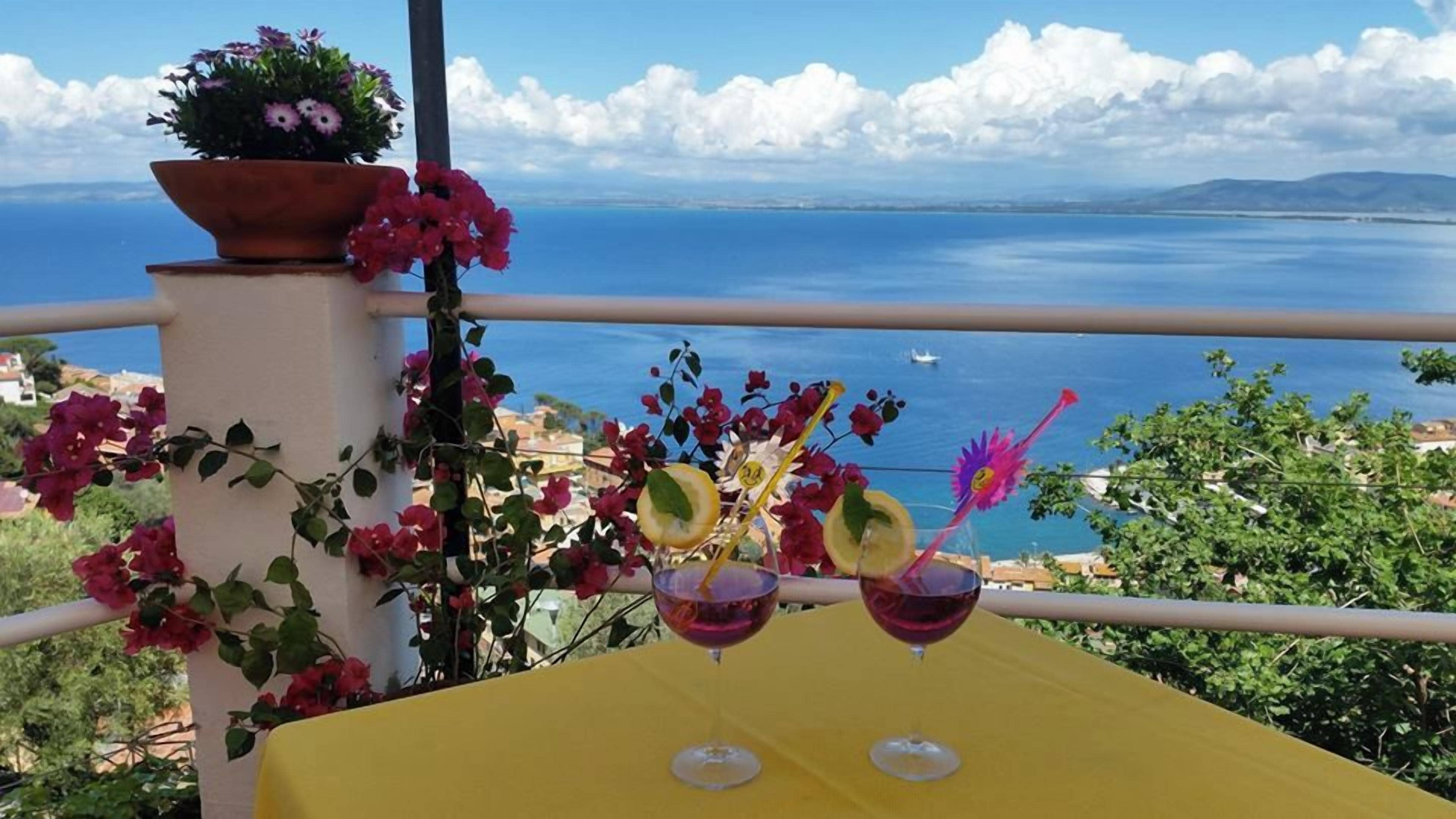A balcony with a yellow table, two glasses of red liquid, and a potted plant with pink flowers.