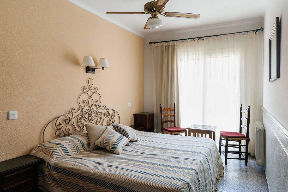 A bedroom with a bed, a chair, a table, a ceiling fan, and a window with curtains.