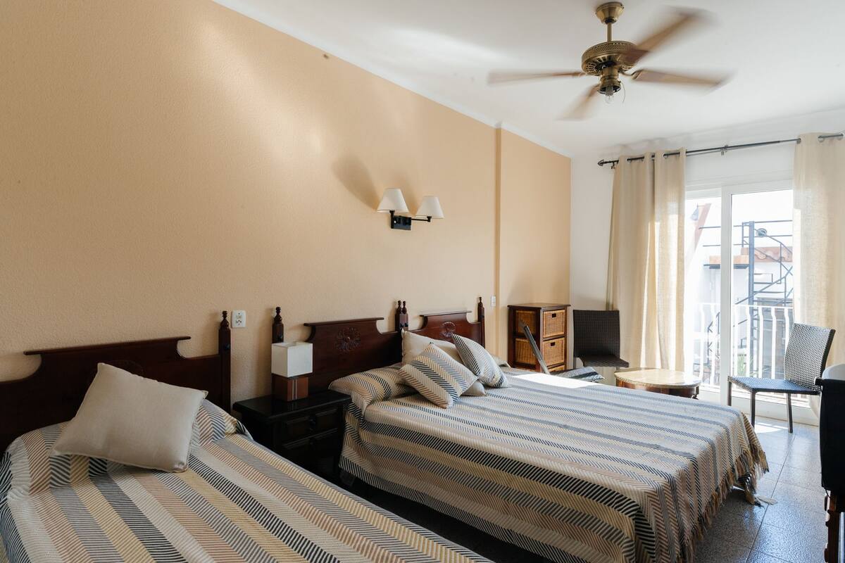 A hotel room with two beds, a ceiling fan, a nightstand, a lamp, a chair, and a view of the outside.