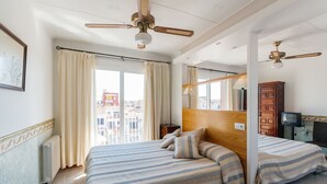 A hotel room with two beds, a ceiling fan, a large window with curtains, a wooden headboard, a television, and a radiator.