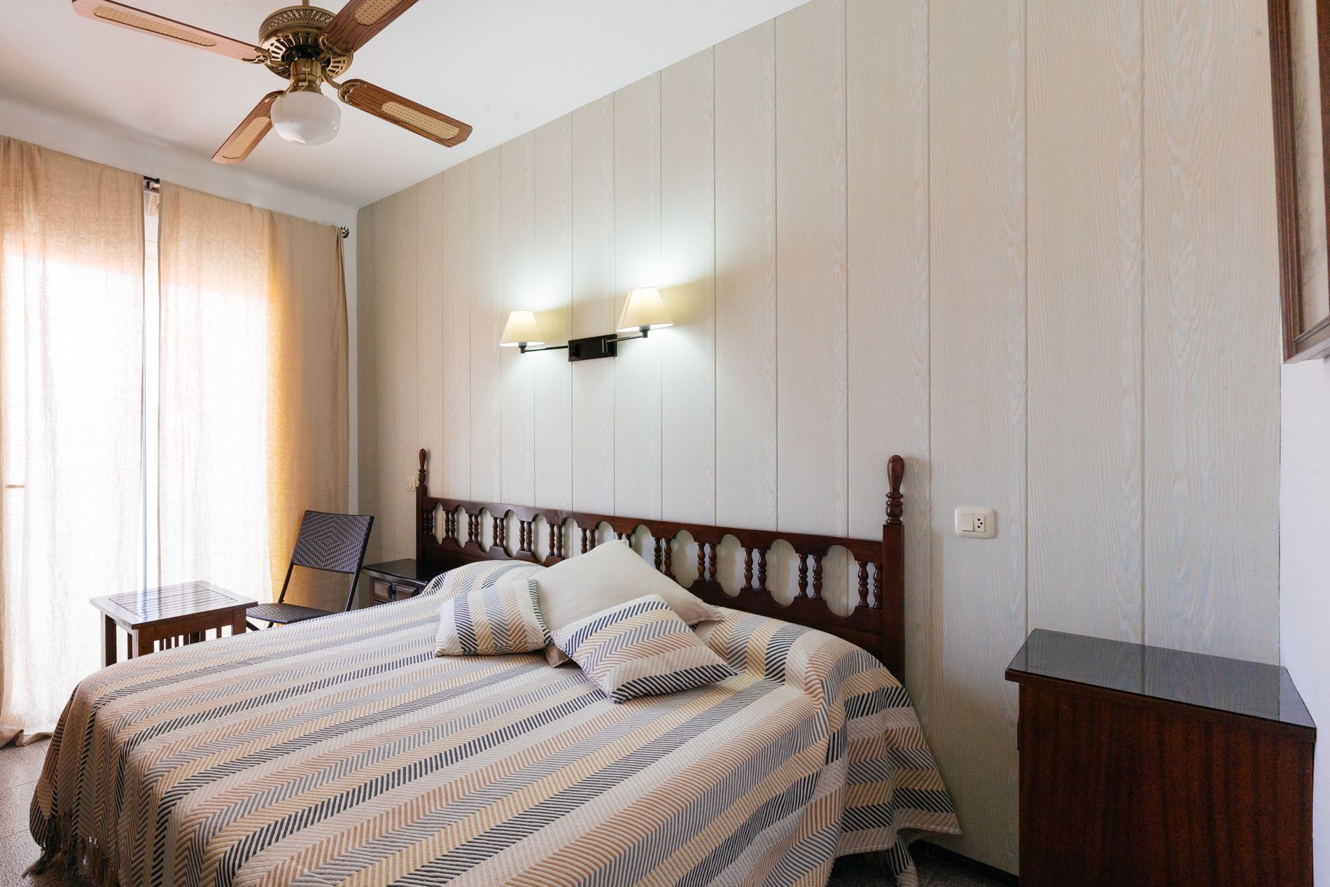 A hotel room with a wooden bed, a desk, a chair, a ceiling fan, and a wall-mounted lamp.