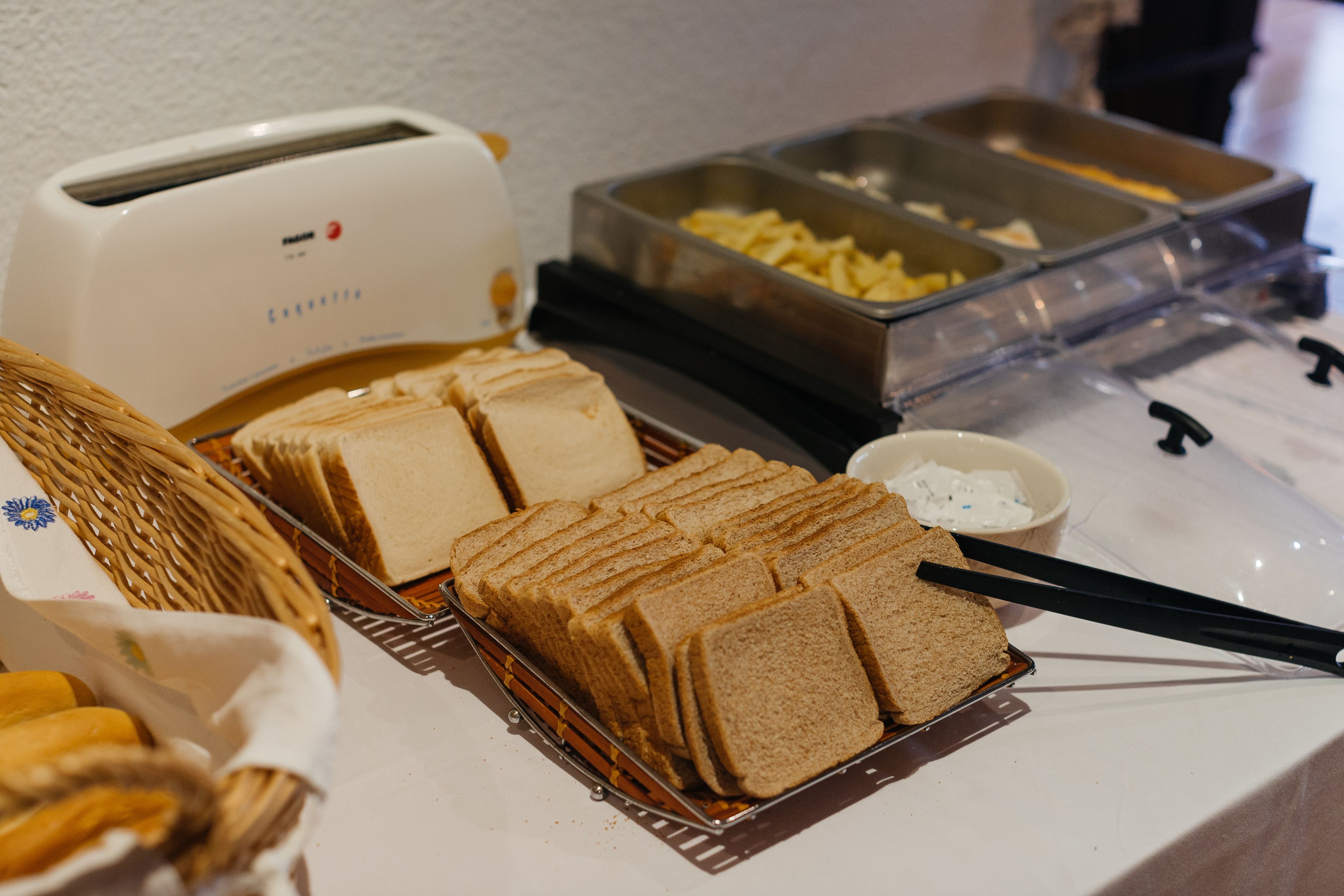 Mesa de buffet com pão fatiado, torradeira e diversos recipientes com comida.