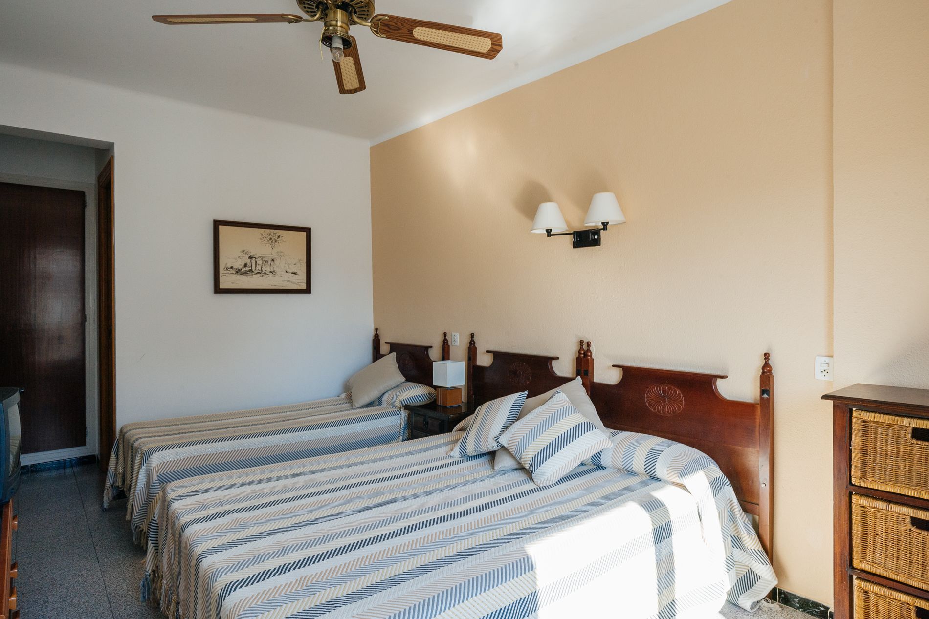 A hotel room with two beds, a ceiling fan, a framed picture on the wall, and a wall-mounted light fixture.
