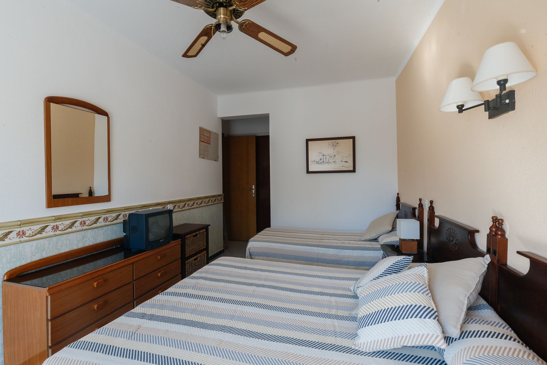 Quarto de hotel com duas camas, um ventilador de teto, cômoda, televisão e luminária fixada na parede.