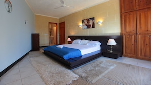 A bedroom with a bed, a ceiling fan, a nightstand, a lamp, and wooden cabinets. - Satori Gozo Centre (Xaghra)