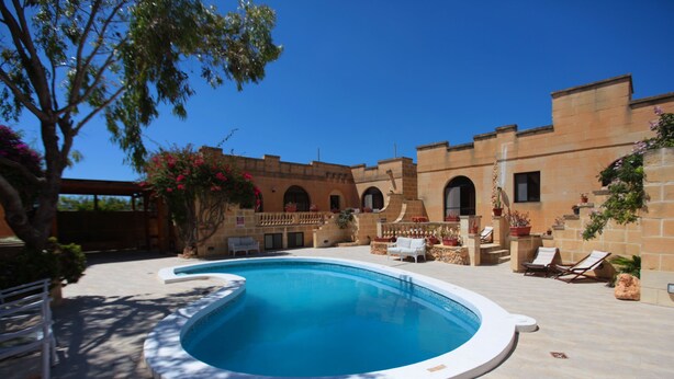 A swimming pool with a curved edge, surrounded by a patio area with lounge chairs and potted plants. - Satori Gozo Centre (Xaghra)