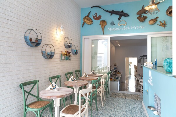 Interior de restaurante con mesas y sillas redondas, una pared azul con decoración temática marina y una entrada visible a otra zona.