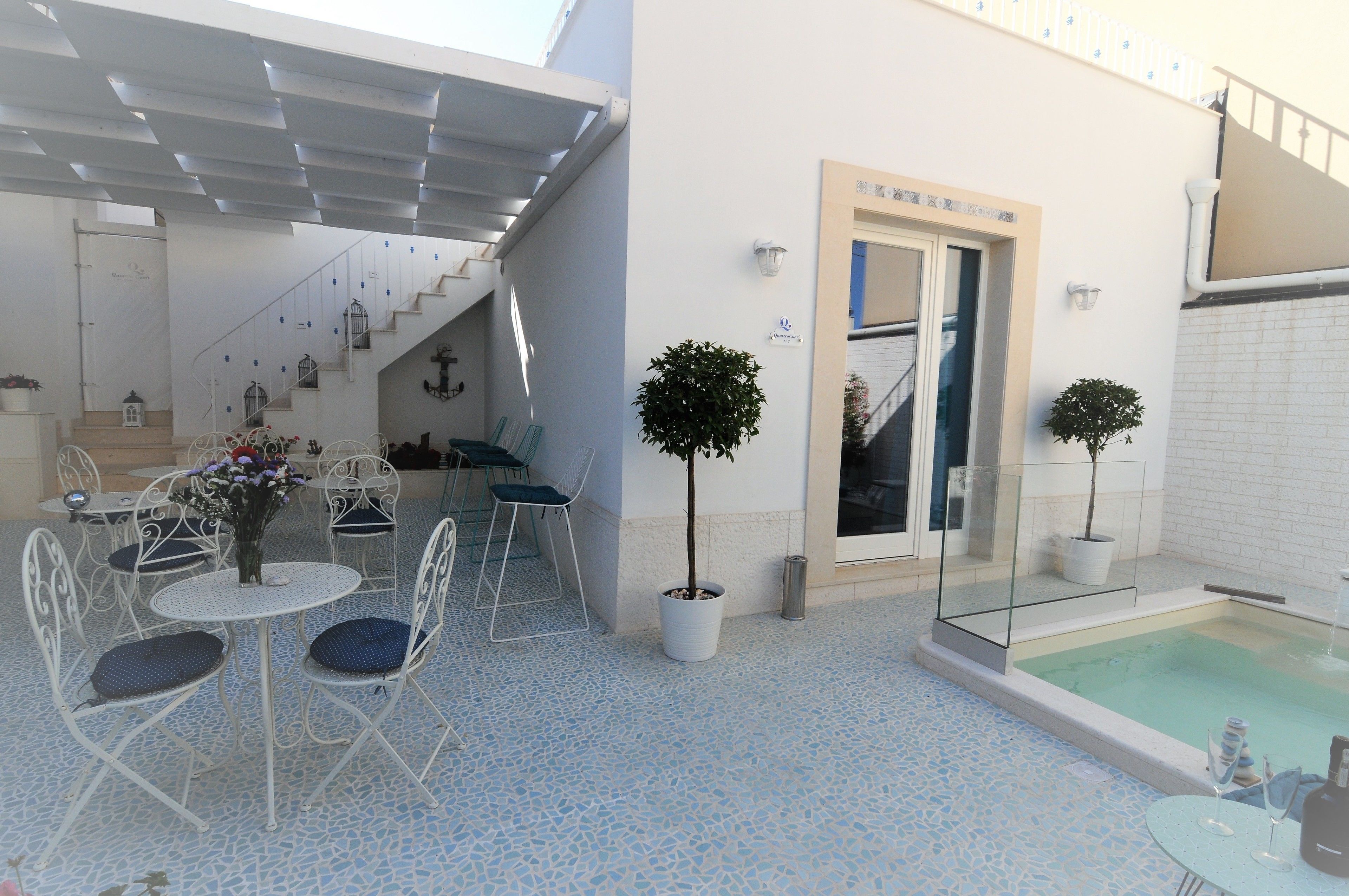 Eine moderne Terrassenanlage mit Pool, weißen Metallstühlen, einem kleinen Tisch mit Blumenvase und einer Treppe, die zu einer oberen Ebene führt.