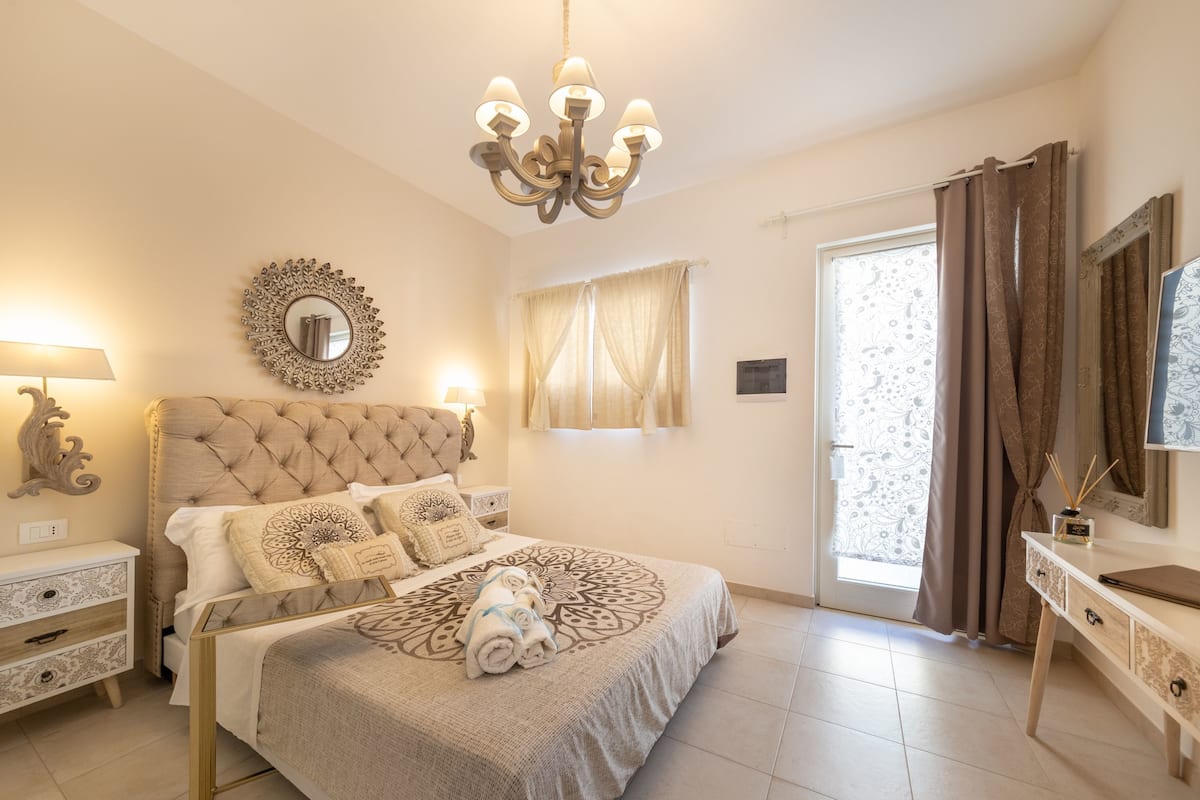 Quarto com cabeceira estofada, cama grande com roupa de cama estampada, lustre, mesa de cabeceira, cômoda e espelho.