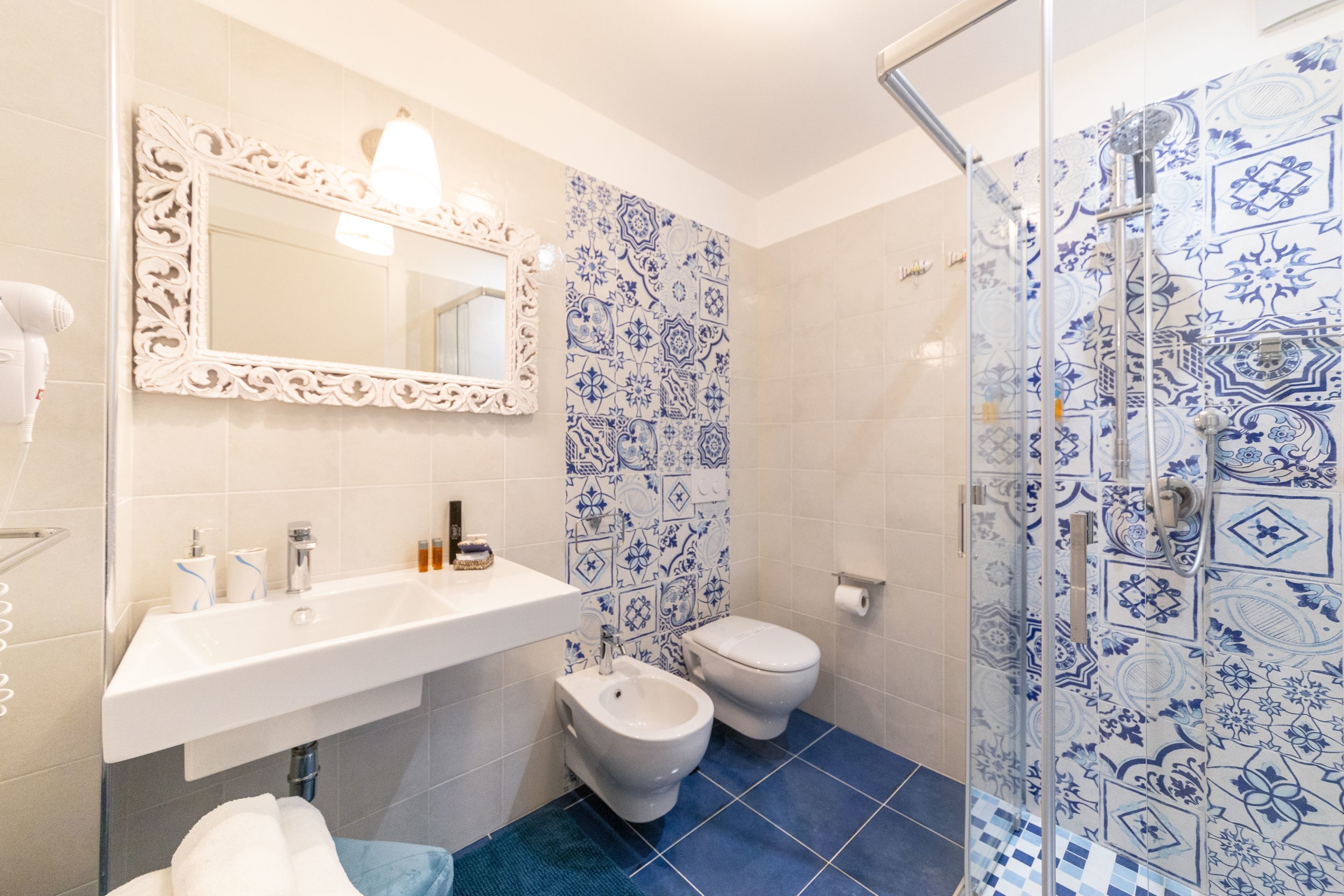 Un bagno con un lavandino bianco, un bidet, un water e una doccia rivestita di piastrelle azzurre a motivi decorativi.