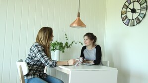 Deux personnes sont assises à une table blanche, au-dessus de laquelle est suspendue une lampe décorative. Une plante en pot se trouve à côté d’elles, et un réveil mural aux chiffres romains est accroché au mur.