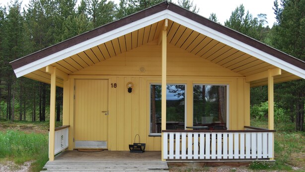 Une cabane jaune avec un porche, une porte portant le numéro 18 et un petit espace porche agrémenté d’une balustrade.