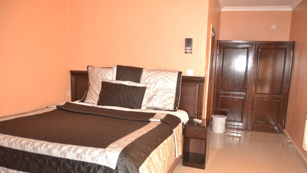 A neatly made bed with a dark and light striped bedspread, a wooden nightstand with a lamp, and a closed door in the background. - Sikalongo Lodge Highlands (Livingstone)