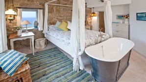 A bedroom with a four-poster bed, a bathtub, a stone wall, and a view of the sea. - Tresillian, Beach House, Sea-views, Large Gardens (Sennen)