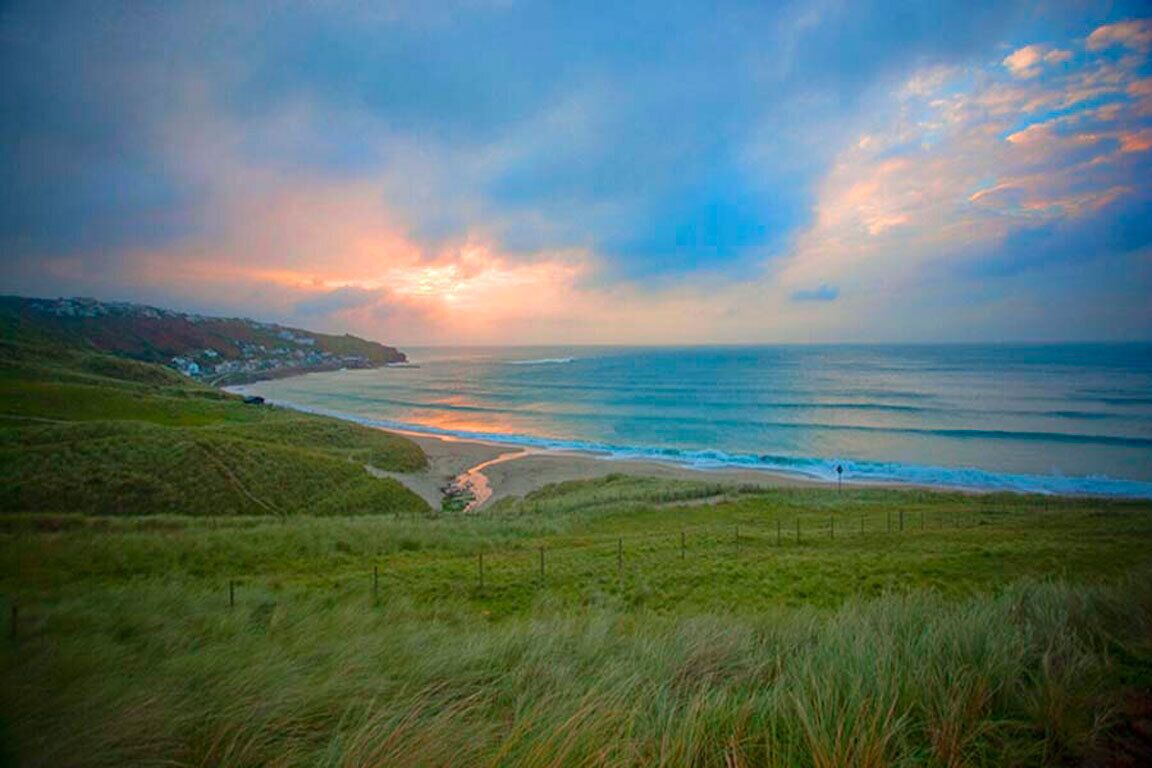 A coastal landscape with a grassy foreground, a beach, and a view of the ocean under a cloudy sky.