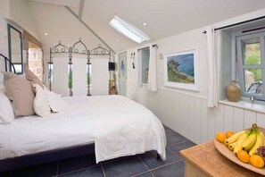 A bedroom with a bed, a window with a view, a wooden table with fruit, and a stone wall. - Basking Shark, Beach Cottage, Superb Sea Views (Sennen)