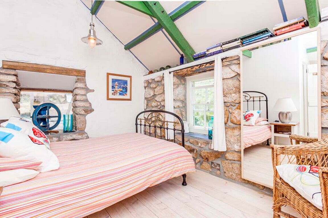 Een slaapkamer met een bed, een rieten stoel, een stenen muur, een raam en een boekenkast.
