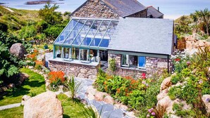 A coastal house with a stone facade, a glass conservatory, and a garden with various plants and flowers. - Castaways, Vellandreath Cornish Cottages (Sennen)