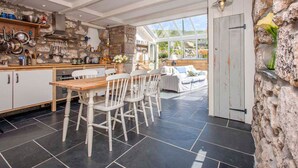 A kitchen with stone walls, a wooden dining table, and a view through a glass door to a garden. - Castaways, Vellandreath Cornish Cottages (Sennen)