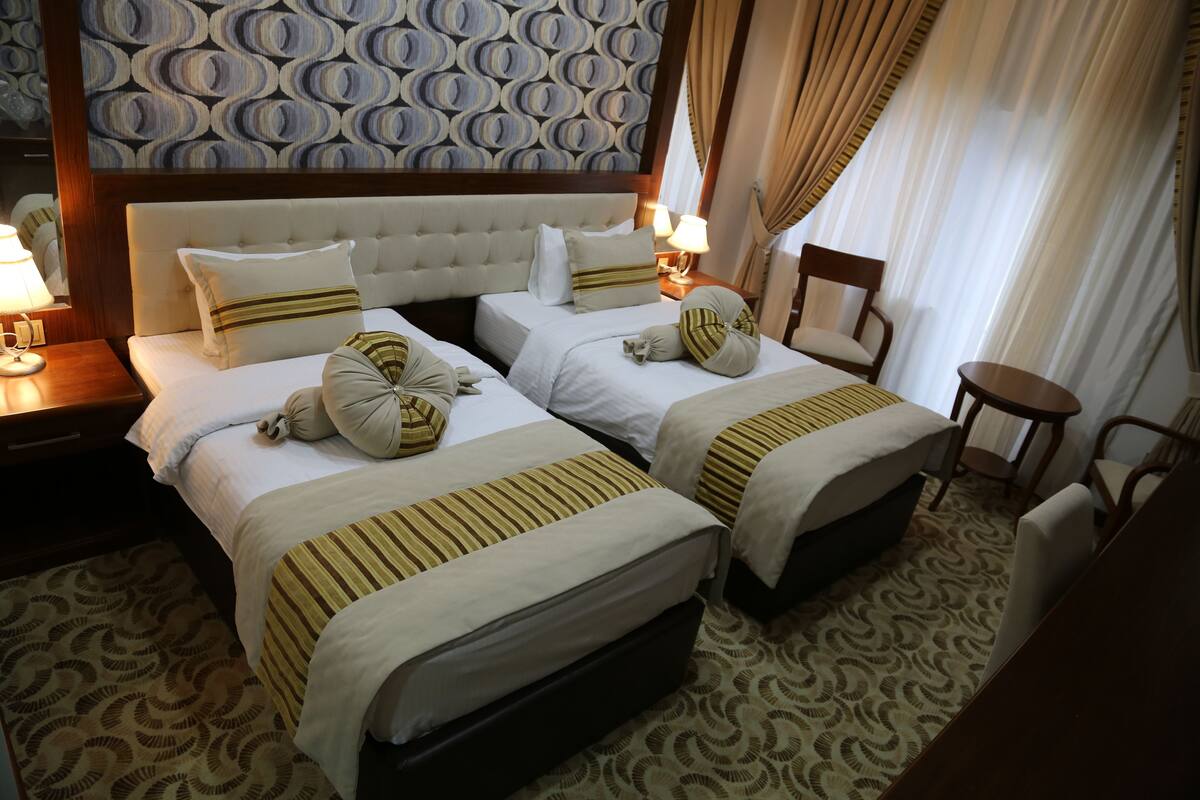 A hotel room with two beds, a headboard, a bedside table with a lamp, a chair, and a patterned carpet.