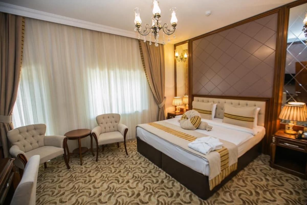 A hotel room with a large bed, two armchairs, a small table, a chandelier, and a large mirror.