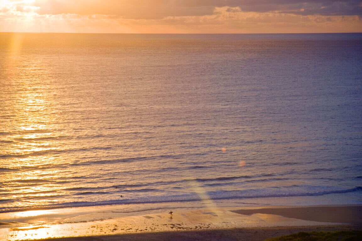 Une plage où l’on voit une personne se promener le long du rivage, l’océan et un coucher de soleil en arrière-plan.