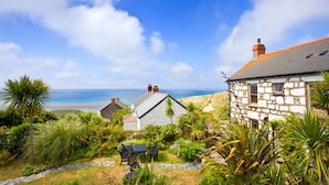 A stone house with a stone roof, surrounded by lush greenery and a view of the sea. - Whitebreakers, Beach Cottage, Sea-views, Garden (Sennen)