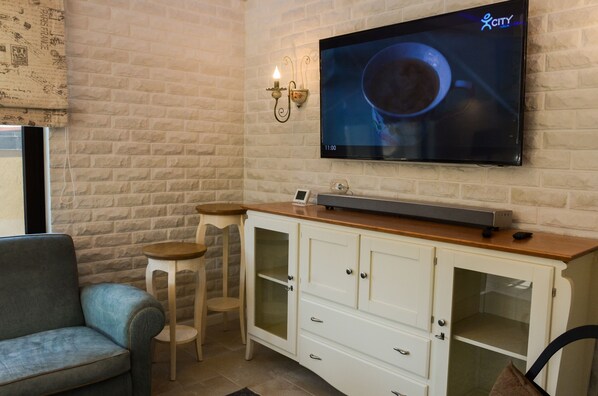 A living room with a flat-screen TV mounted on a brick wall, a white cabinet with drawers, a blue sofa, and two wooden stools. - Residence Bilyana (Svilengrad)